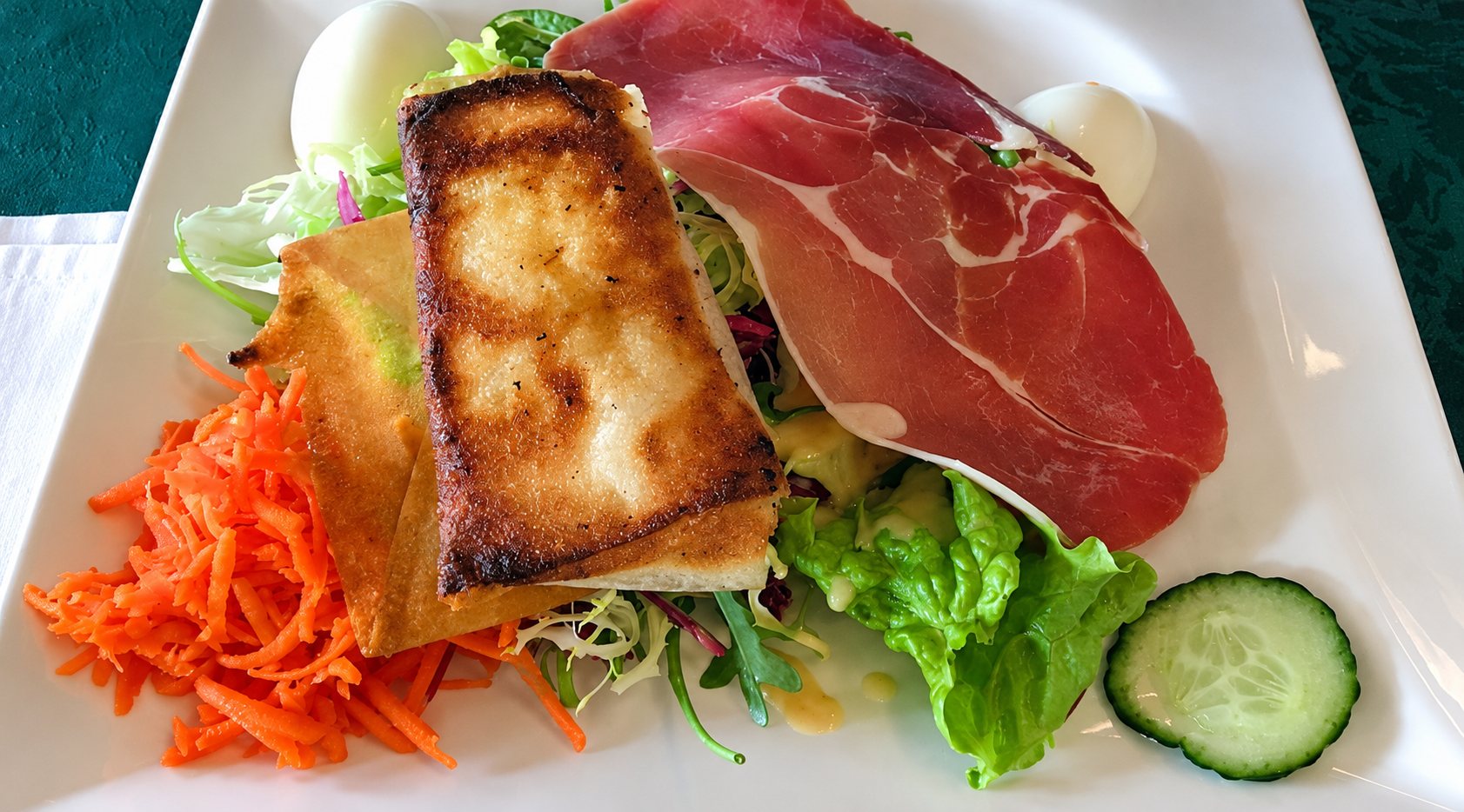 Assiette carrée blanche garnie de salade verte, carottes râpées, jambon cru, œufs durs, concombre et feuilleté doré.