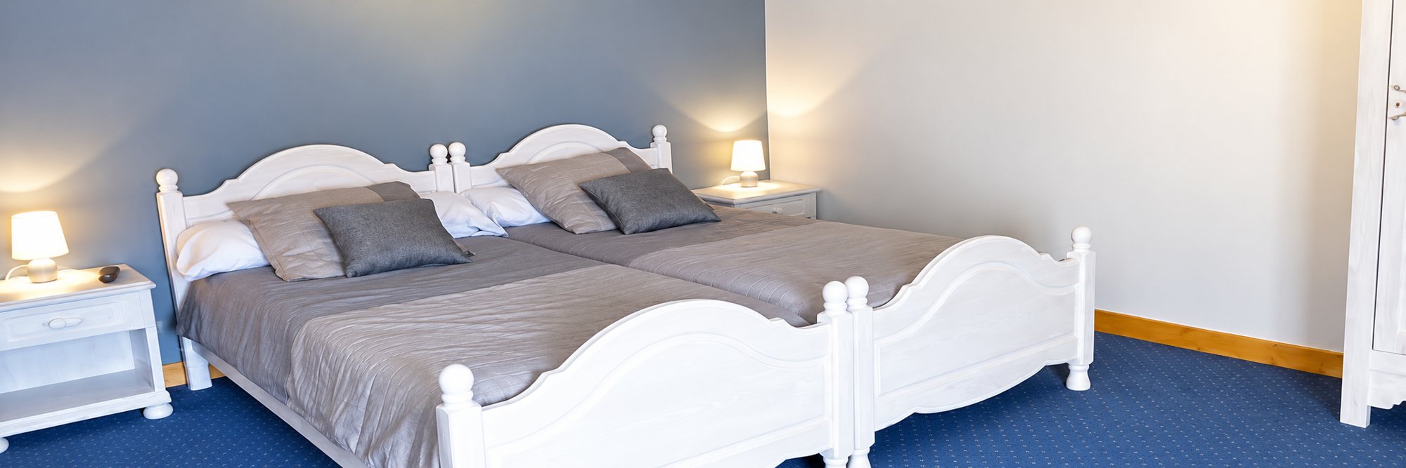 Chambre aux murs bleu-gris avec deux lits blancs en bois jumelés, couvertures grises, deux lampes de chevet allumées et moquette bleue.