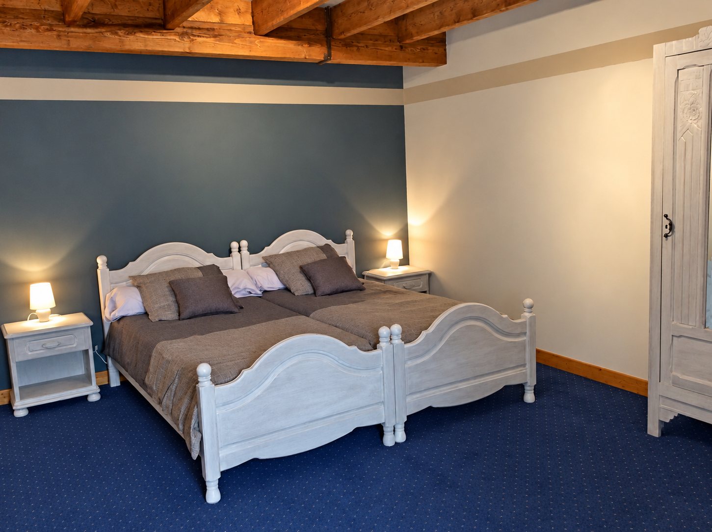 Chambre d'hôtes avec deux lits jumeaux en bois blanc, deux tables de chevet éclairées, un mur bleu-gris et des poutres au plafond.