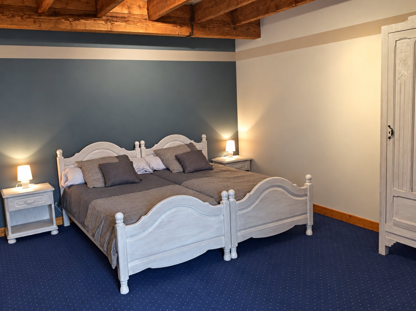 Chambre d'hôtes avec deux lits en bois blanc, deux tables de chevet éclairées, une armoire ancienne, des poutres apparentes et une moquette bleu foncé.
