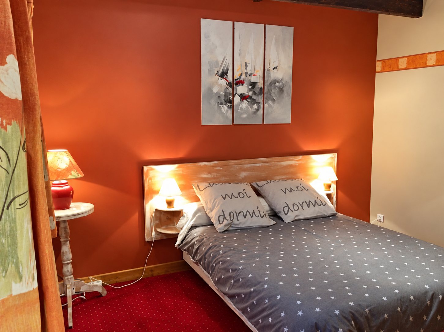 Chambre aux murs orange terracotta avec un lit à couette grise étoilée, deux coussins imprimés de texte, une tête de lit éclairée et un tableau triptyque.