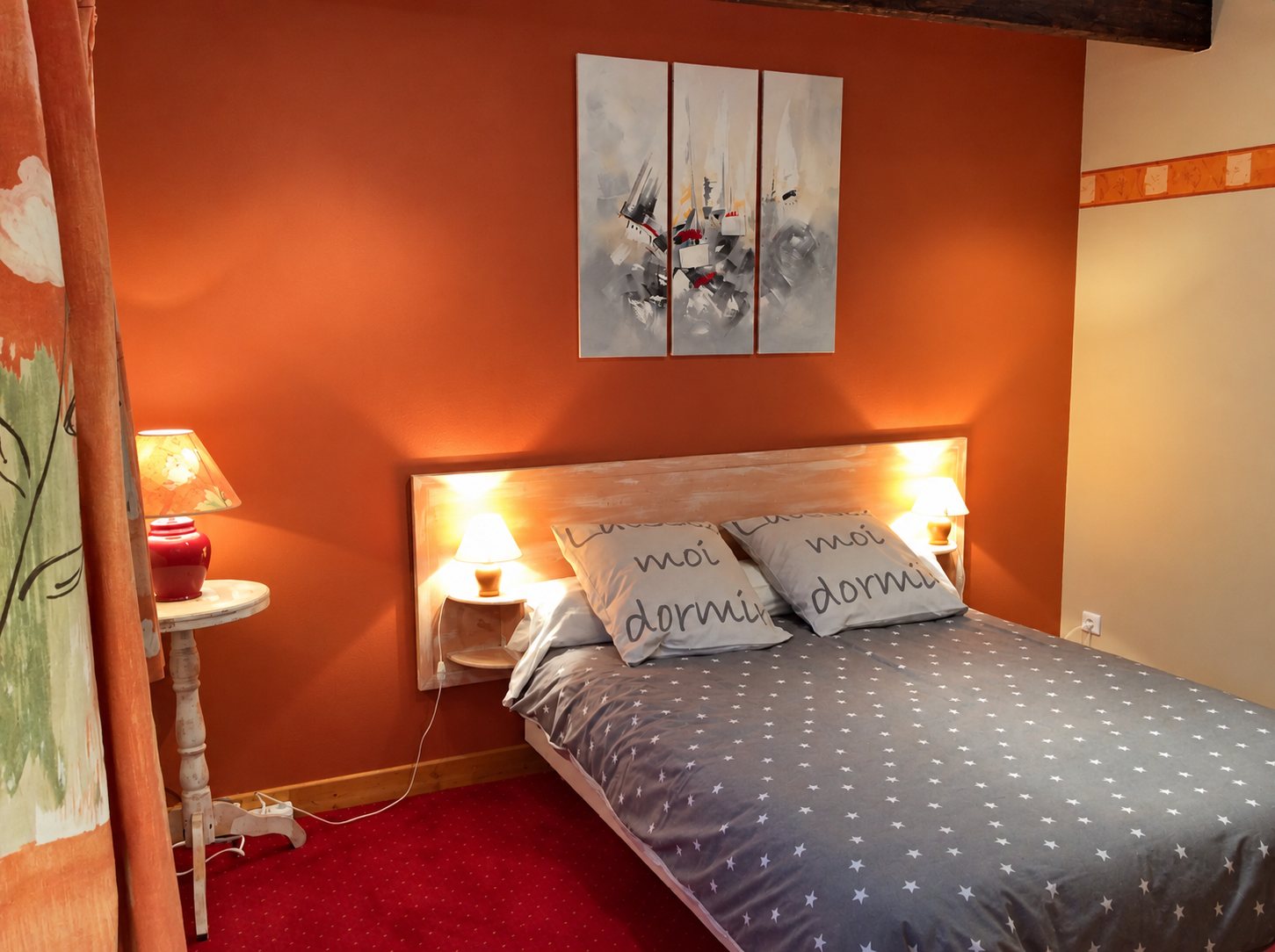Chambre aux murs orange terracotta avec un lit double à housse grise étoilée, deux coussins imprimés, des lampes allumées et un triptyque abstrait au mur.