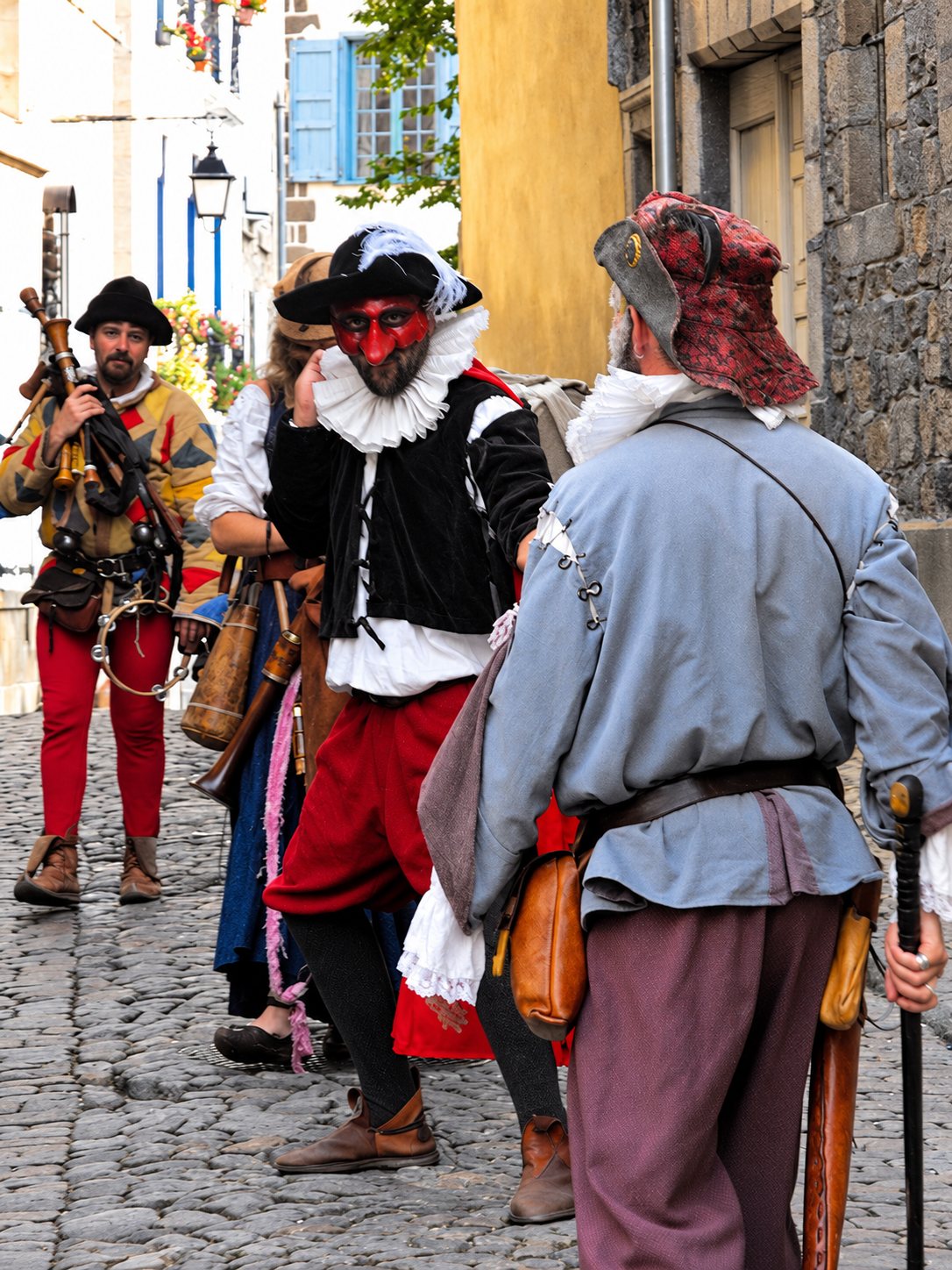 Un groupe de personnes en costumes d'époque, certaines au visage masqué de rouge, discutent dans une ruelle pavée bordée de façades anciennes.