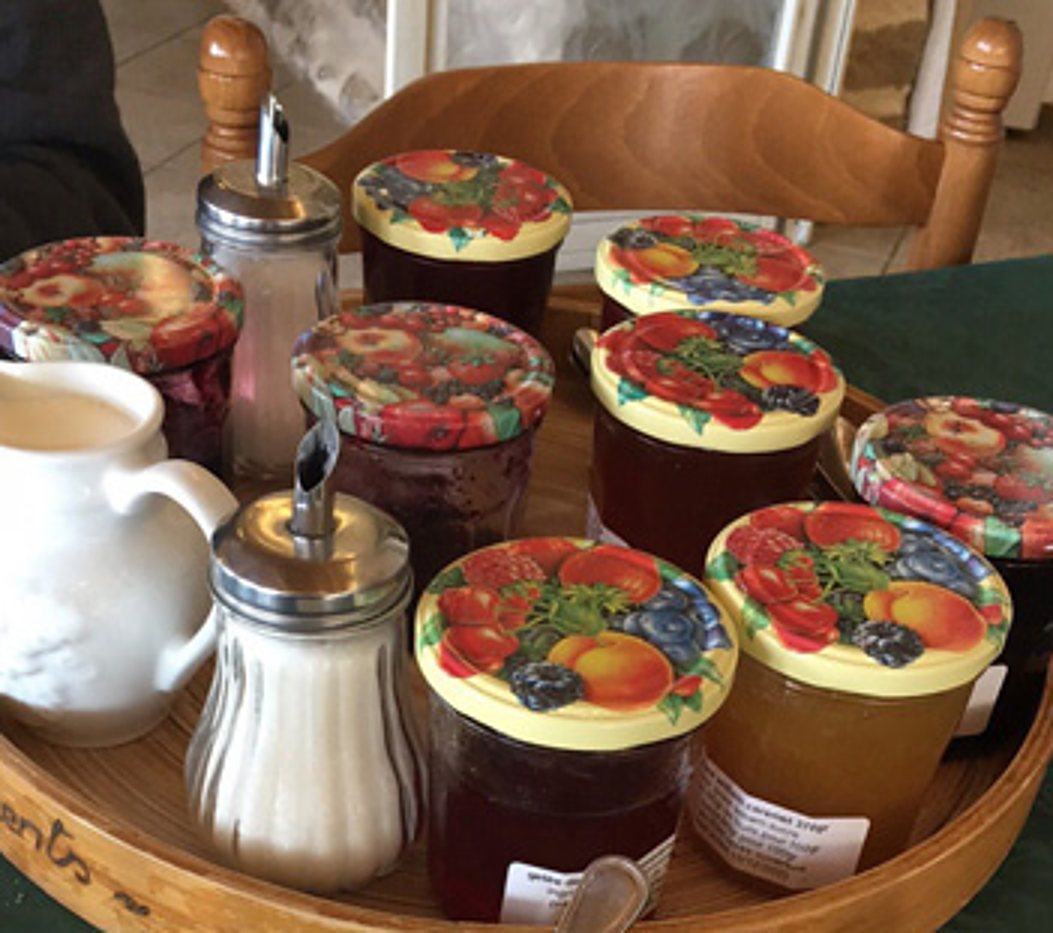 Un plateau rond en bois posé sur une table verte porte plusieurs pots de confiture aux couvercles à motif de fruits, un sucrier verseur en verre et un pichet blanc.