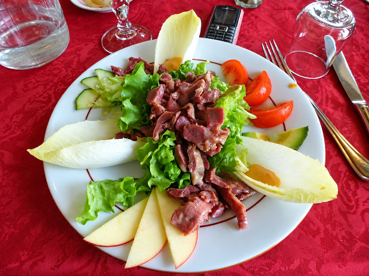 Assiette de salade composée avec gésiers, endives, salade verte, tomates, concombre, avocat et quartiers de pomme, posée sur une nappe rouge.