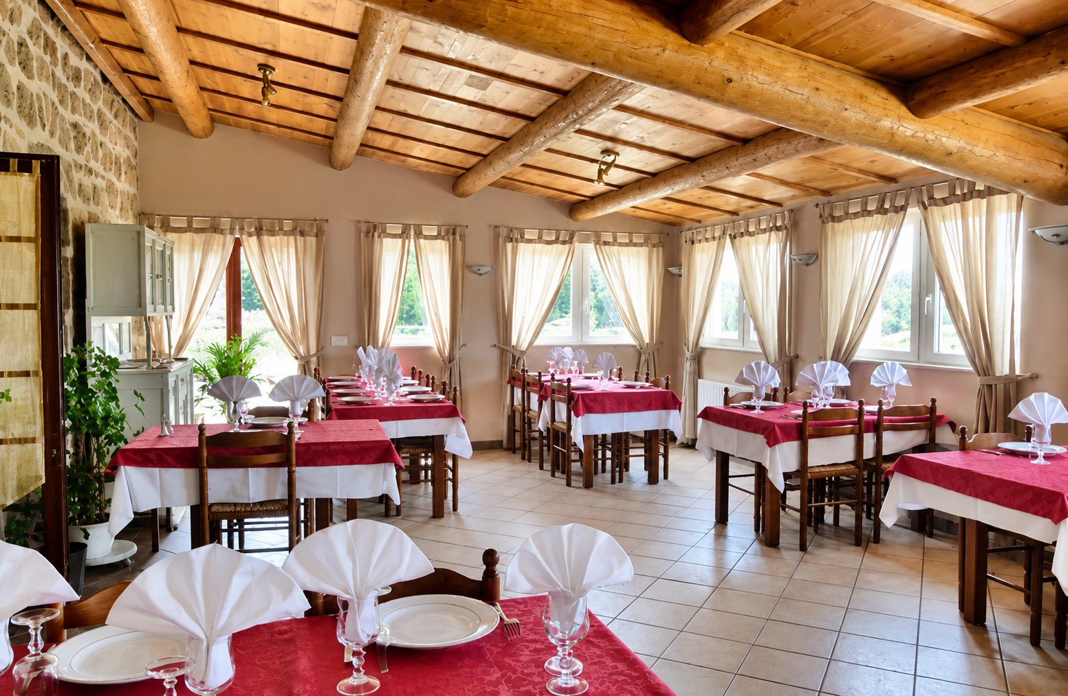Salle de restaurant lumineuse aux poutres en bois et au mur de pierre, avec des tables dressées de nappes rouges et blanches et de serviettes pliées.