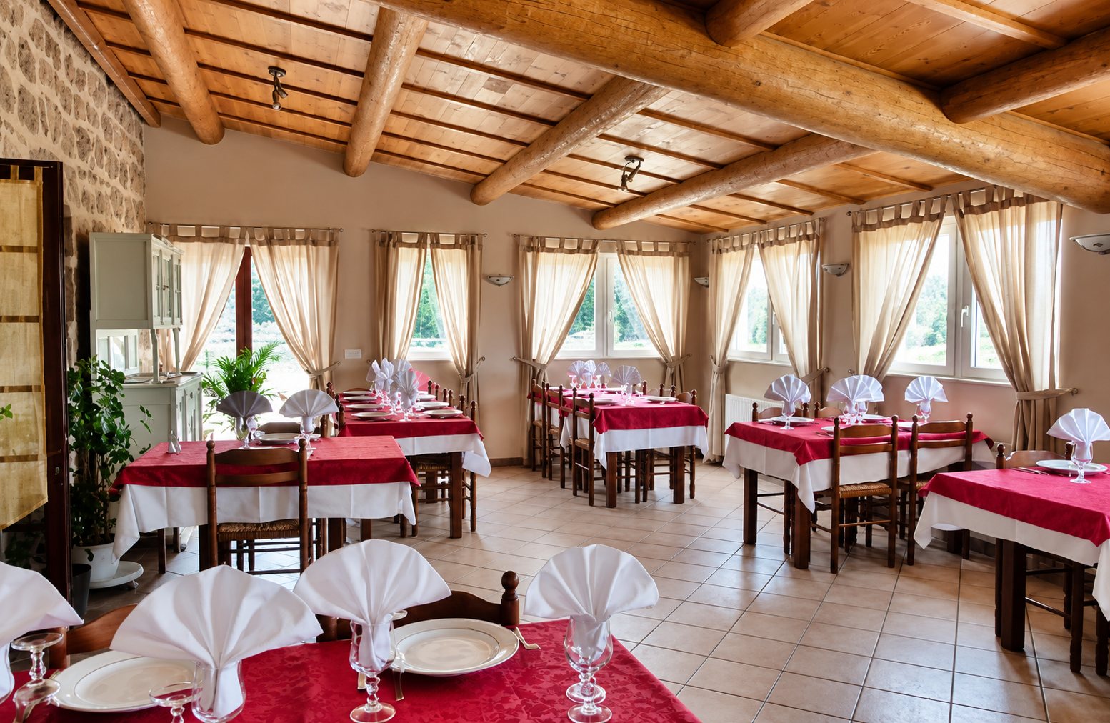 Salle de restaurant lumineuse aux poutres en bois et murs en pierre, avec des tables dressées de nappes rouges et blanches et de serviettes pliées en éventail.