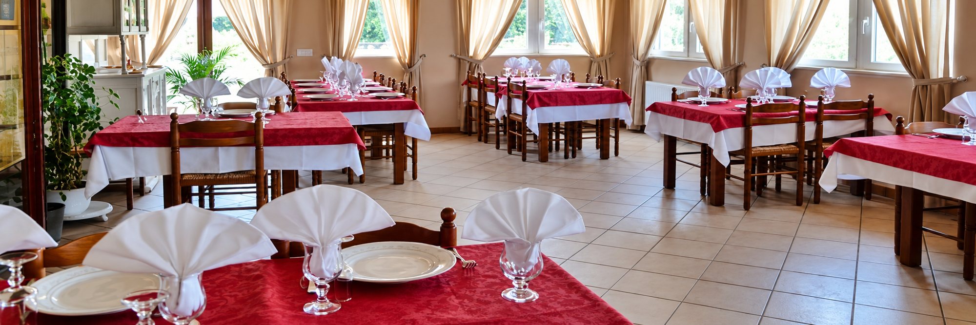 Salle de restaurant lumineuse avec des tables en bois dressées de nappes rouges et blanches, d'assiettes, de verres et de serviettes pliées en cornet.