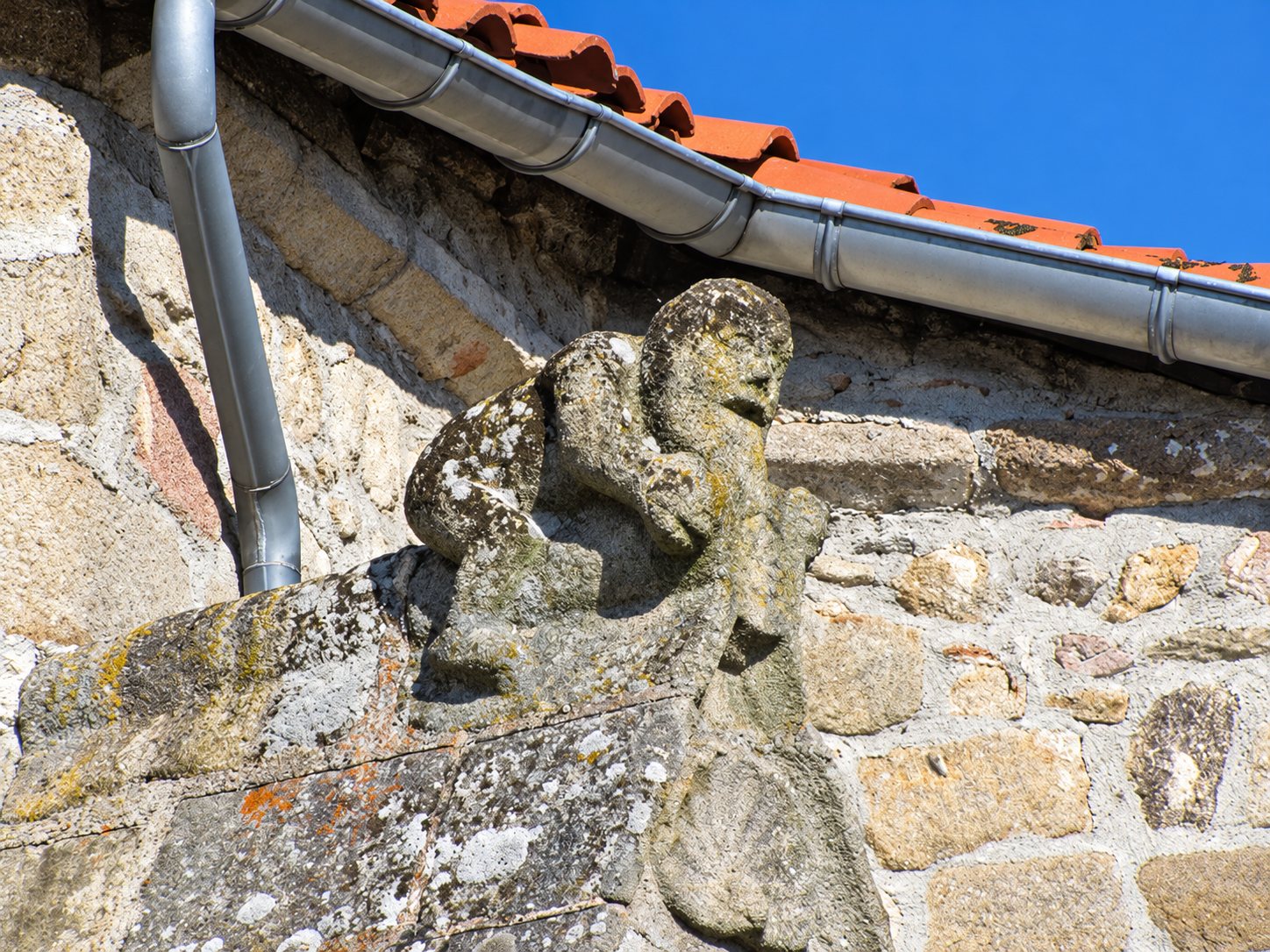 Une figure sculptée en pierre couverte de lichen est fixée sur un mur en pierres, sous une gouttière métallique et un toit de tuiles rouges.